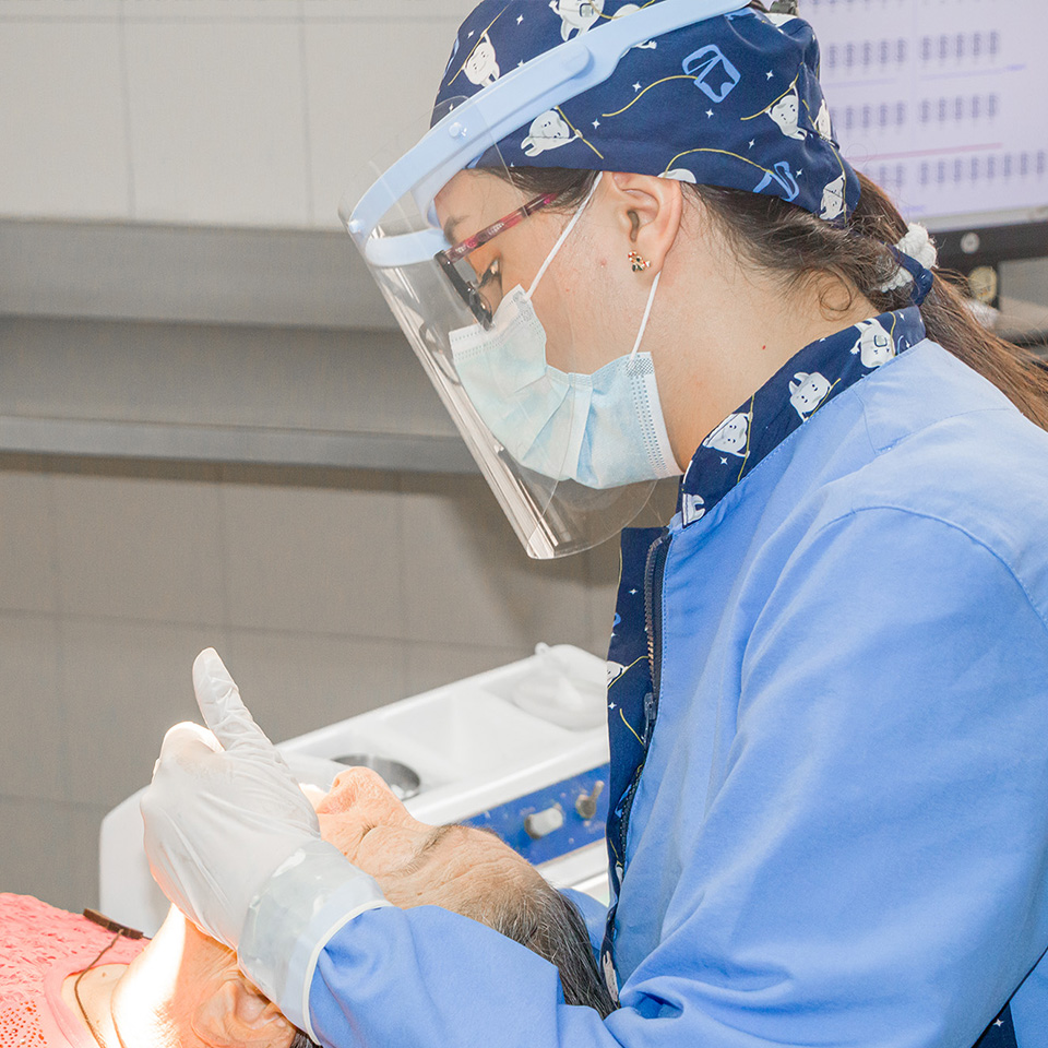 Odontóloga con mascarilla de protección revisa a un paciente acostado, sosteniendo instrumentos dentales para la revisión.